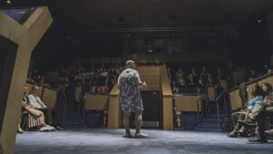 A person standing in a large auditorium, talking to a crowd of speakers